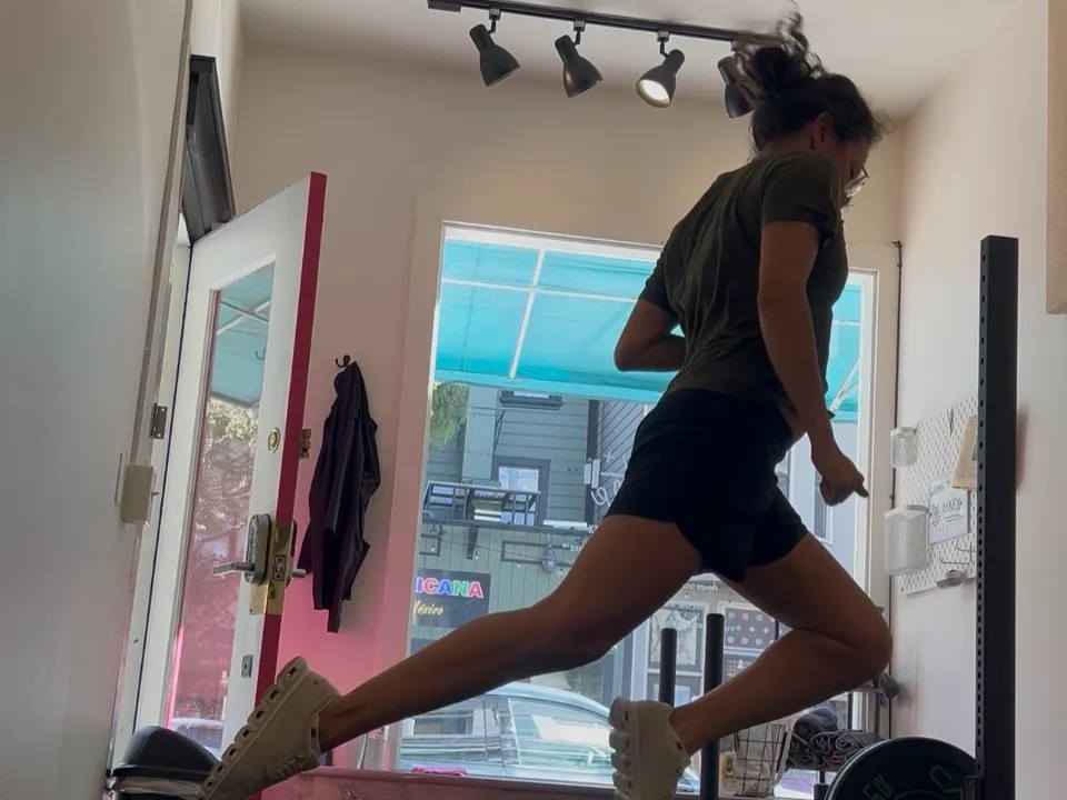 Zulma mid-air in a jumping lunge in a small training studio